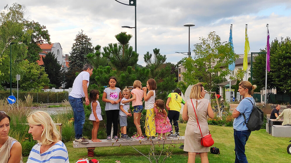 Das Bildzeigt den neu errichteten Grünraum mit spielenden Kindern und Erwachsenen, die sich unterhalten