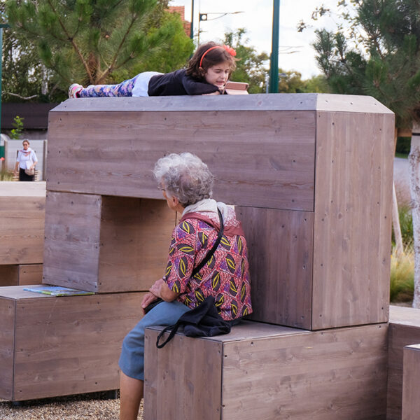 ältere Dame sitzt auf Holzskulptur, kleines Mädchen liegt oben und schaut auf Handy