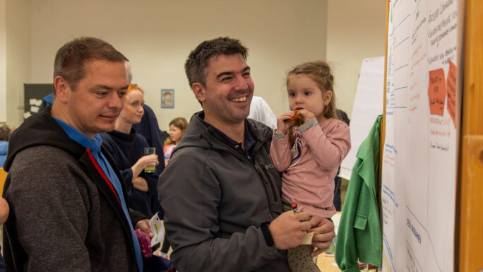 zwei junge Männer, einer davon mit Kind am Arm, diskutieren an einem Flipchart