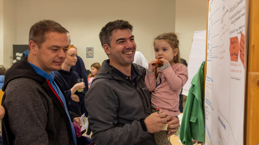 Zwei Männer, einer mit Kind auf auf dem Arm, diskutieren vor einem Flipchart