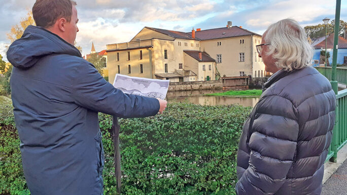 Das Foto zeigt zwei Männer, die auf dem Grünraum vor einem öffentlichen Gebäude auf einen Plan schauen.