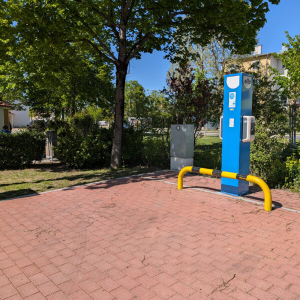 Eine blaue E-Ladestation, davor ein rot gepflasterter Platz, dahinter Bäume und eine grüne Wiese