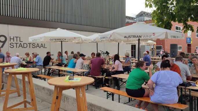 Menschen sitzen bei einem FEst an Tischen, sonnenschirme sind auch zu sehen