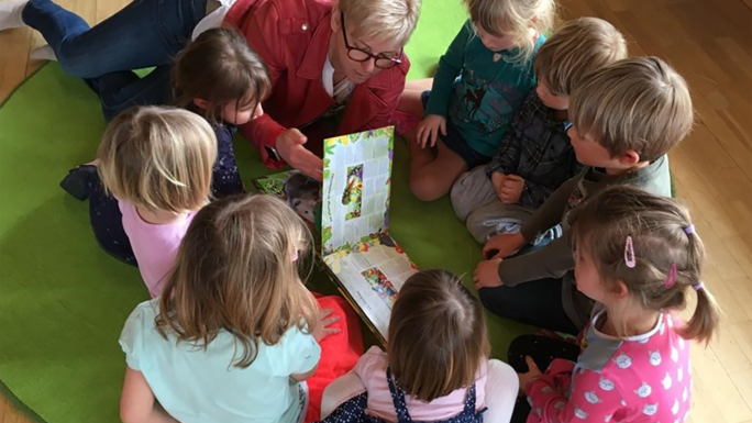 Acht Kinder, die am Boden sitzend gemeinsam mit einer Betreuerin ein Buch ansehen