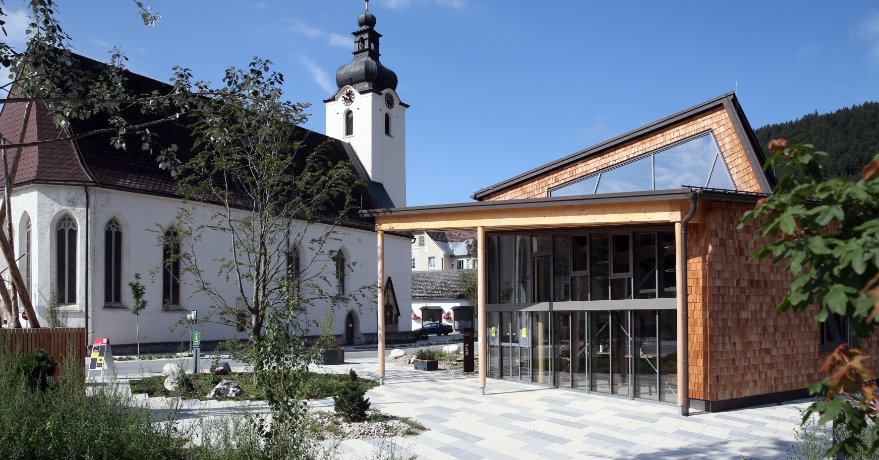 Kirche im Hintergrund, im Vordergrund modernes Holzgebäude und gepflegter Vorplatz