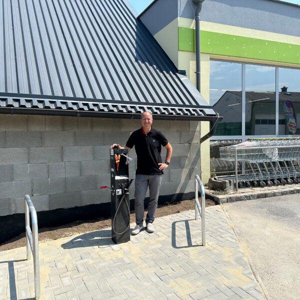 ein Mann steht bei einer Radreparatursäule, dahinter ist der Supermarkt zu sehen
