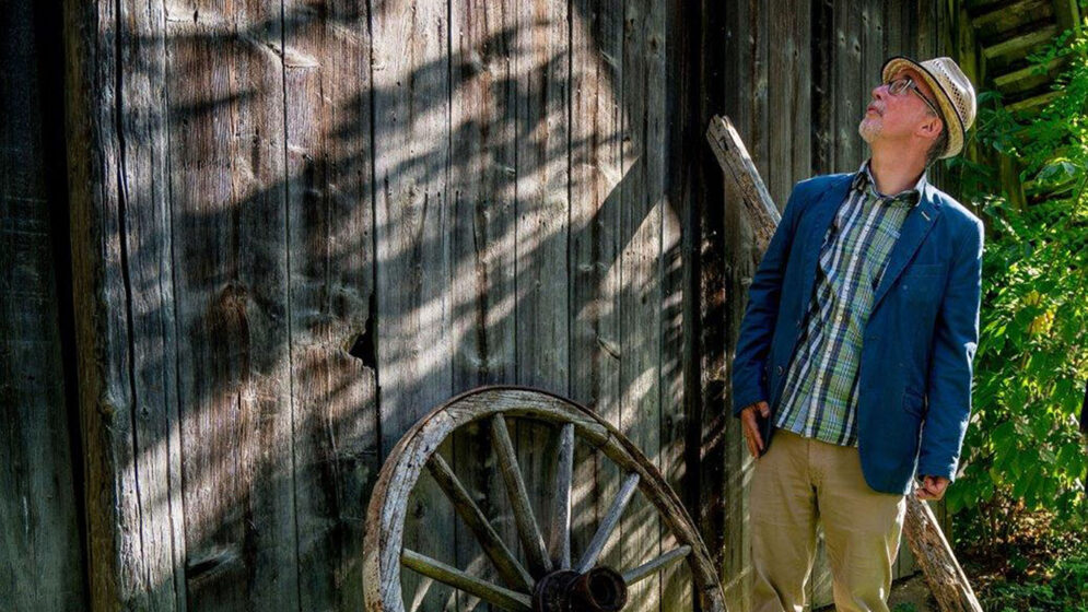 Ein Mann schaut auf einen Stadel, an dem ein altes Holzrad lehnt.