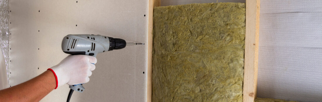 Nahaufnahme einer Arbeiterhand mit elektrischem Schraubendreher beim Befestigen von Trockenbauplatten an einem Holzständerwerk mit Dämmung aus Mineralwolle. Warmes Zuhause, niedrige Energiekosten, Konzept für Bau und Renovierung.