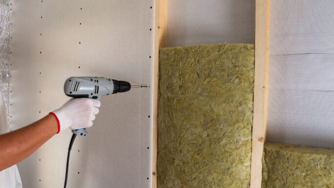 Nahaufnahme einer Arbeiterhand mit elektrischem Schraubendreher beim Befestigen von Trockenbauplatten an einem Holzständerwerk mit Dämmung aus Mineralwolle. Warmes Zuhause, niedrige Energiekosten, Konzept für Bau und Renovierung.