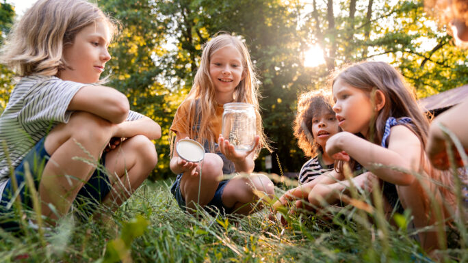ein Mädchen sitzt in der Wiese und hält in einer Hand ein Glas und in der anderen den Deckel dazu. Drei Kinder liegen dabei und sehen ihr zu.