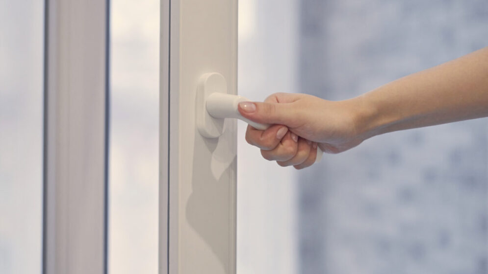 Frau hält einen weißen modernen Griff an einem PVC-Fenster mit Doppelverglasung und öffnet bzw. schließt den Fensterflügel. Nahaufnahme.﻿
