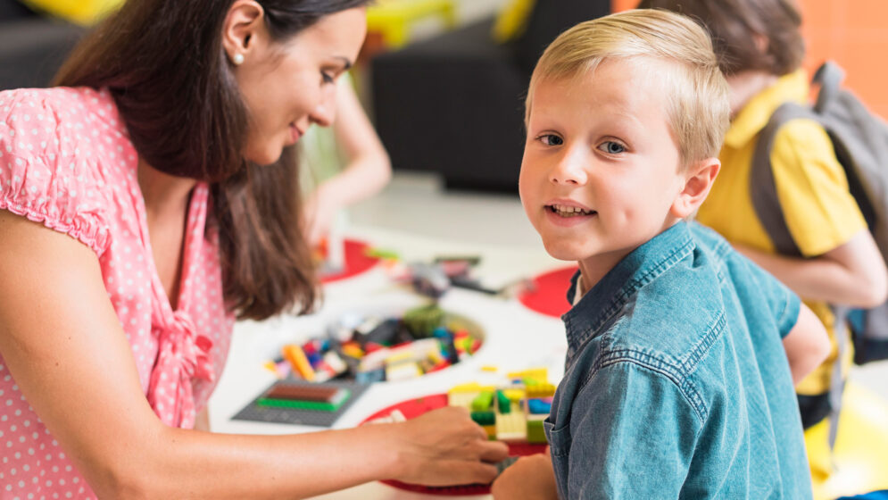 Lehrerin spielt mit Kindern an einem Tisch, ein Junge im Vordergrund schaut direkt in die Kamera