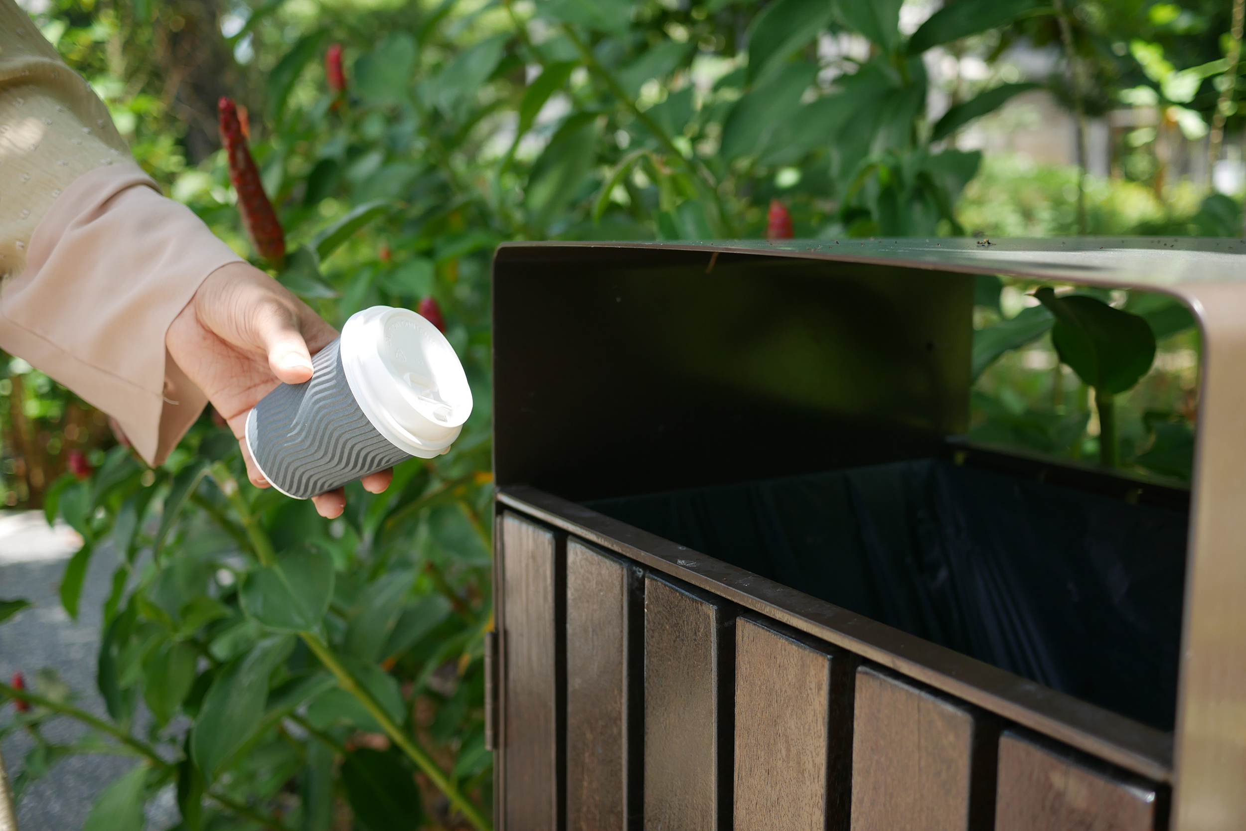 eine Person wirft einen gebrauchten Kaffeebecher in einen Mistkübel im Freien